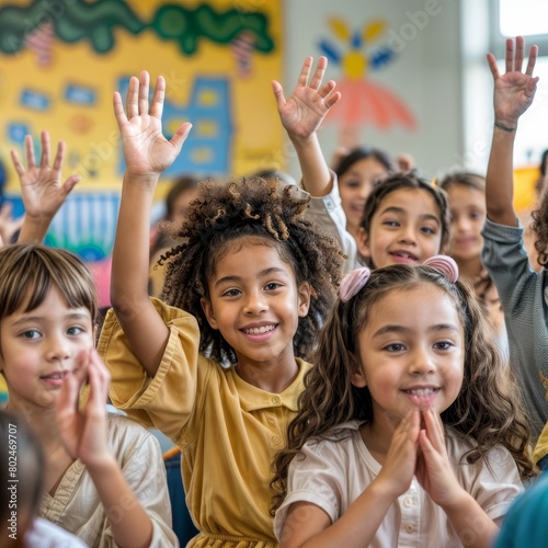 A group of children are in a classroom, with one girl in a yellow shirt and another girl in a white shirt. They are all smiling and raising their hands in the air. Scene is happy and positive