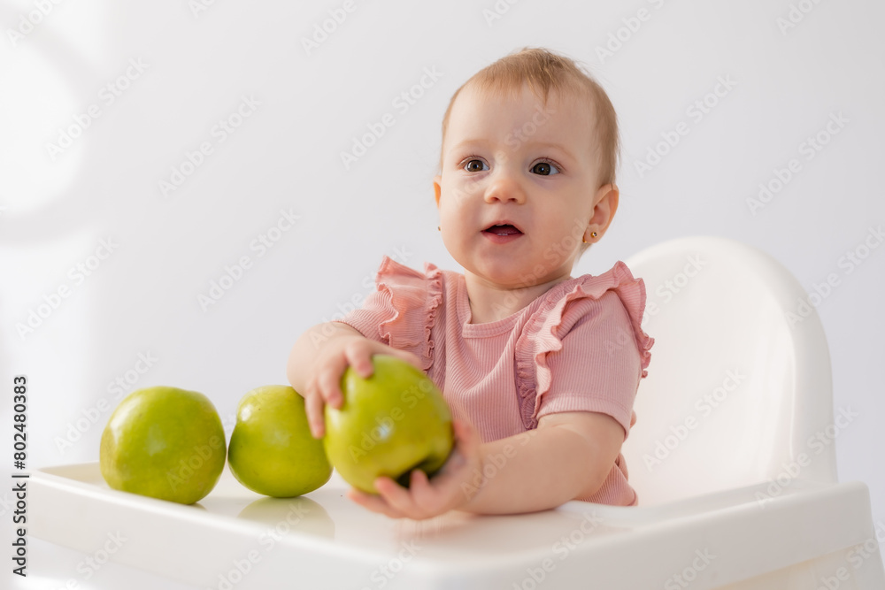 Cute baby in a baby chair nibbles apples on a white background Cute baby in a baby chair nibbles apples on a white background