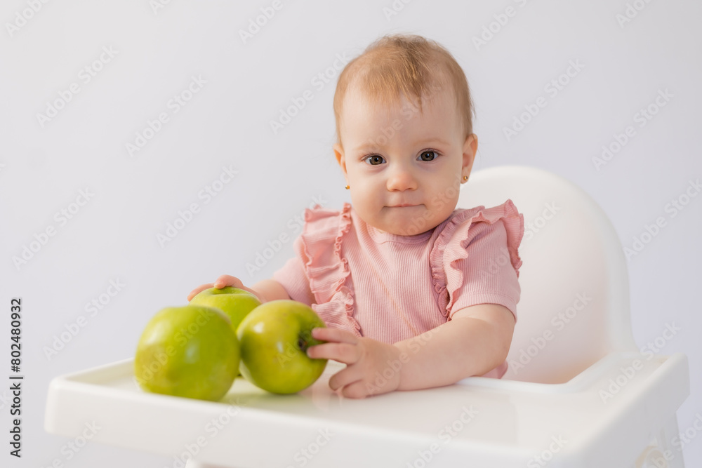 Cute baby in a baby chair nibbles apples on a white background Cute baby in a baby chair nibbles apples on a white background