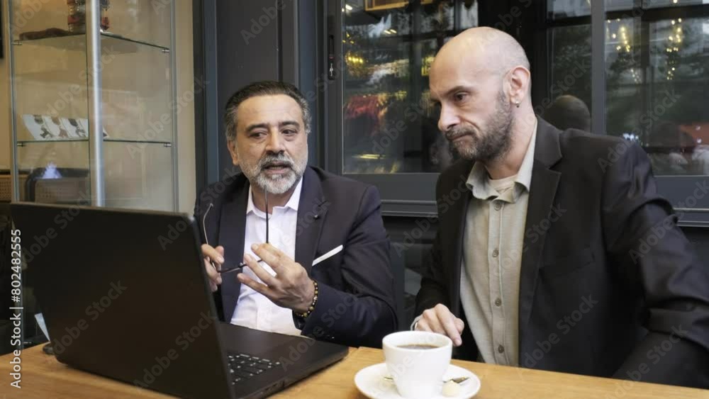 Two charismatic middle-aged businessmen, dressed in suits, are having a meeting outside the office in a cafe environment, laughing heartily while working with their laptops Two charismatic middle-aged businessmen, dressed in suits, are having a meeting outside the office in a cafe environment, laughing heartily while working with their laptops