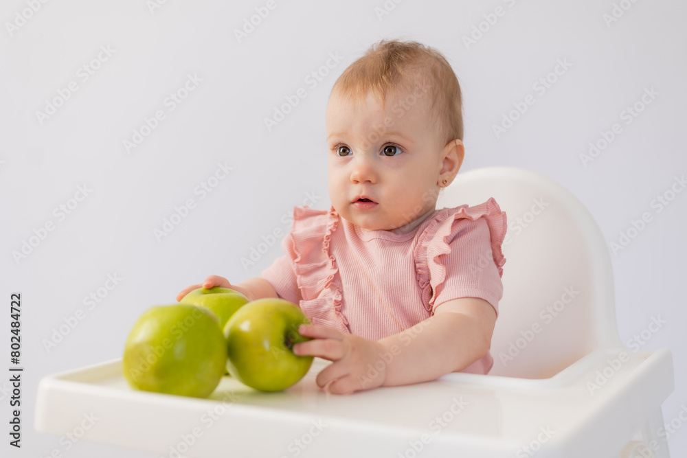 Cute baby in a baby chair nibbles apples on a white background Cute baby in a baby chair nibbles apples on a white background