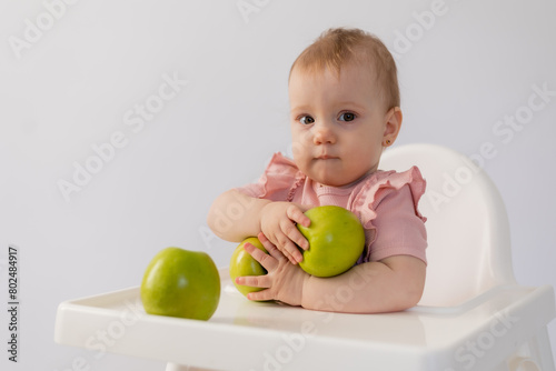 Cute baby in a baby chair nibbles apples on a white background