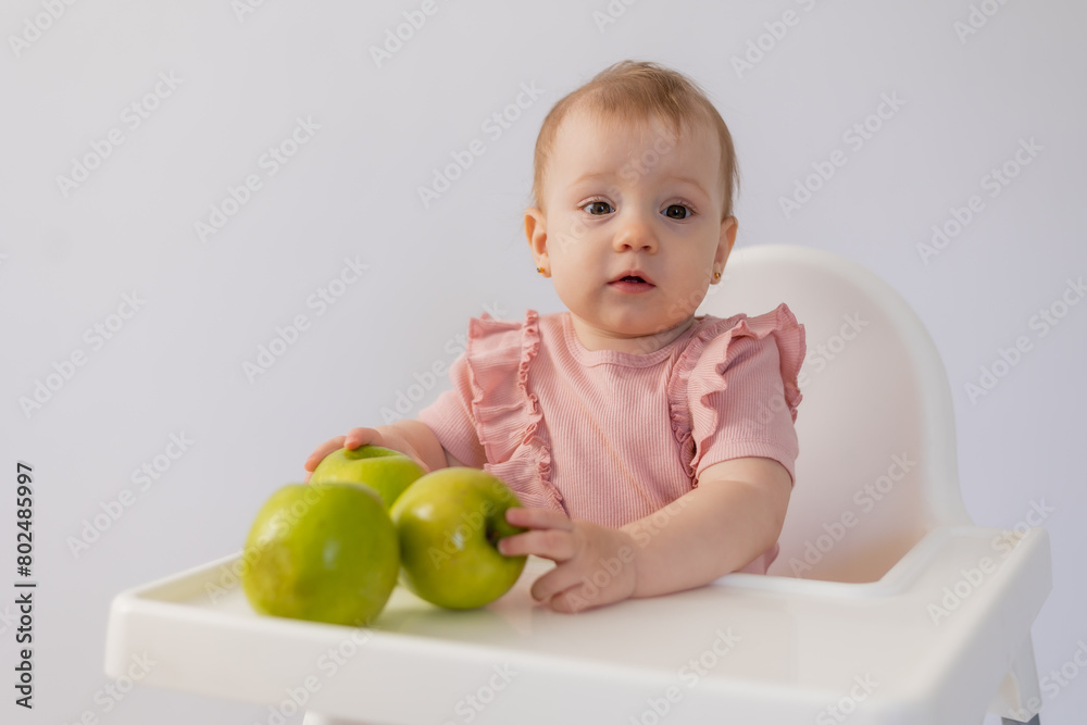 Cute baby in a baby chair nibbles apples on a white background Cute baby in a baby chair nibbles apples on a white background
