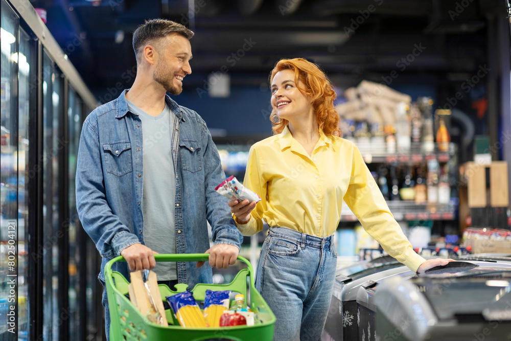 Happy young couple shopping together at supermarket, husband and wife standing by freezer with shopping trolley full of food