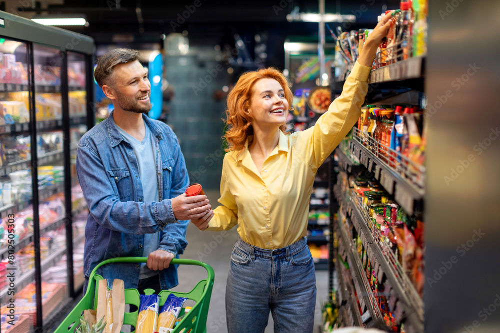 Smiling spouses shopping in hypermarket, choosing and buying food, walking with trolley cart in supermarket indoors