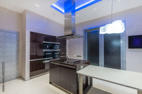 Stylish kitchen with light walls and dark furniture. Glass door and bright blue ceiling .lighting.