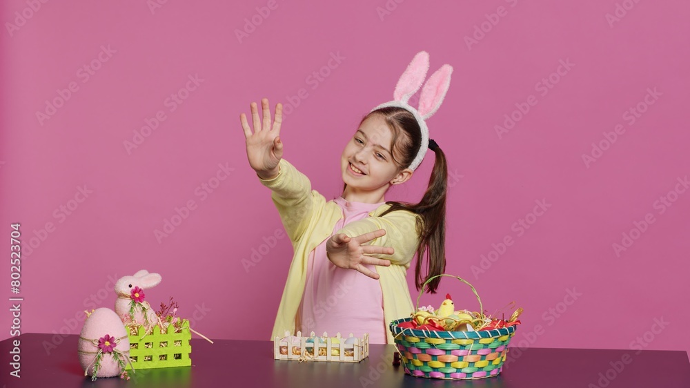Energetic young girl with adorable bunny ears waving in studio, saying hello and greeting someone while she creates easter decorations. Joyful toddler posing against pink backdrop. Camera A.