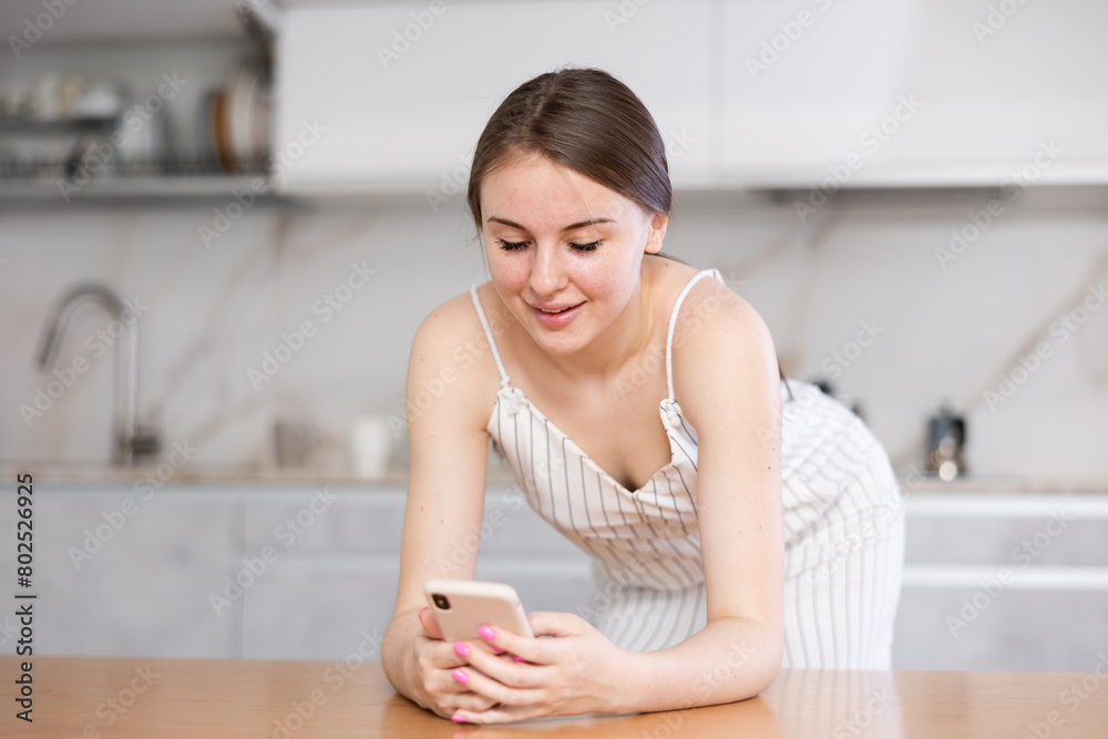 Teen girl leaning on table and watching something interesting on her mobile phone in the kitchen