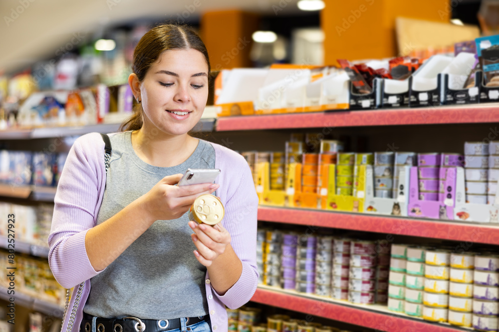 Interested young woman using smartphone to scan ingredients label on small tin can while choosing healthy organic nutritious wet food