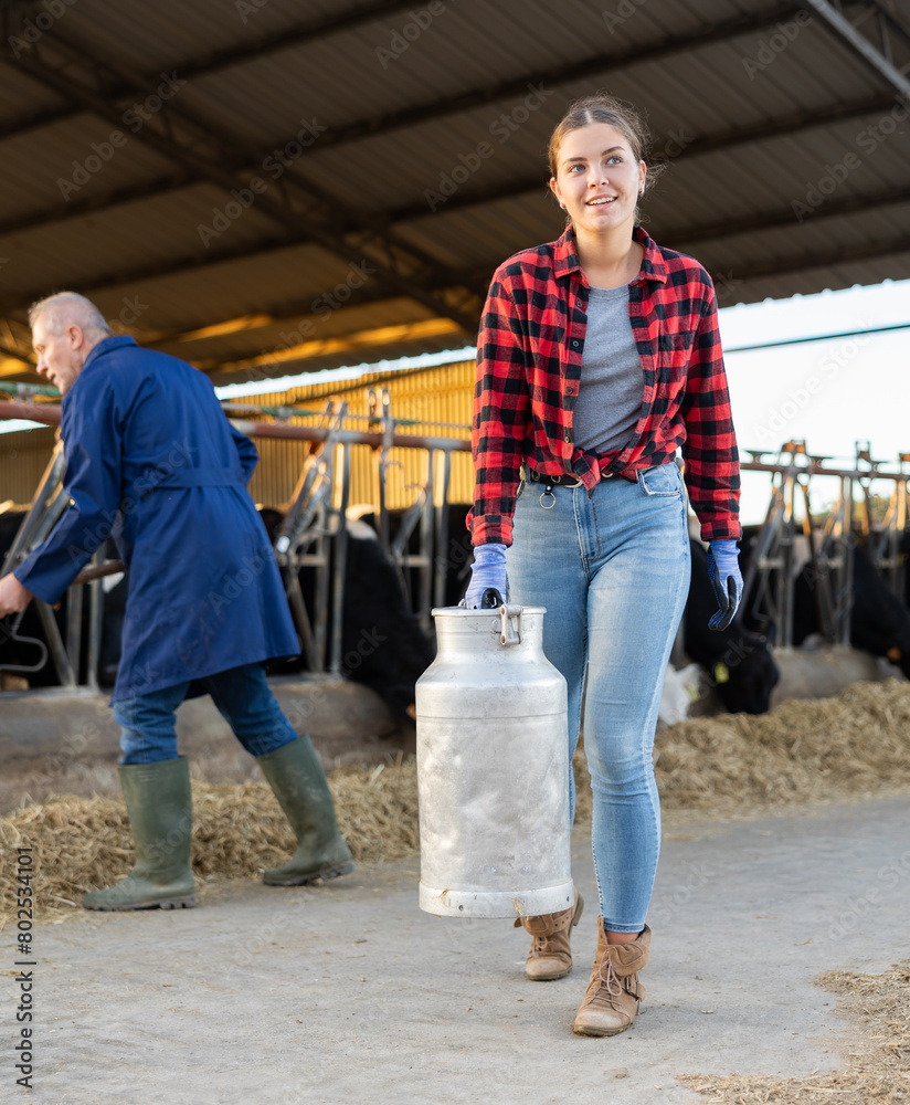 Positive female farmer with large metallic milk can in hangar with cows