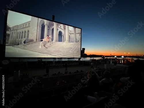 An open-air cinema, offering romantic leisure and entertainment on summer evenings with a high-quality screen and film. People enjoying a movie at sunset by the river.