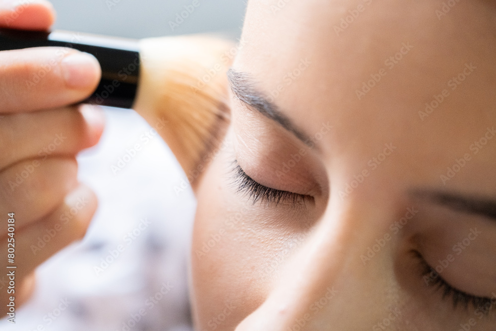 Detail of a young teenager applying blush to her cheeks in a relaxing manner with a soft brush. The light is soft, and the eye remains completely focused.