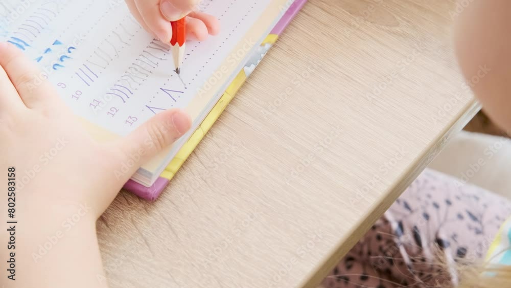 5-year-old girl diligently draws elements with pencil in notebook on table close-up, learning to write, spiral college block, student writing supplies, back to school