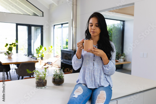 Young biracial woman sitting at home, enjoying coffee in bright kitchen photo