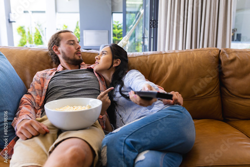 Diverse couple relaxing on couch at home, watching TV and eating popcorn photo