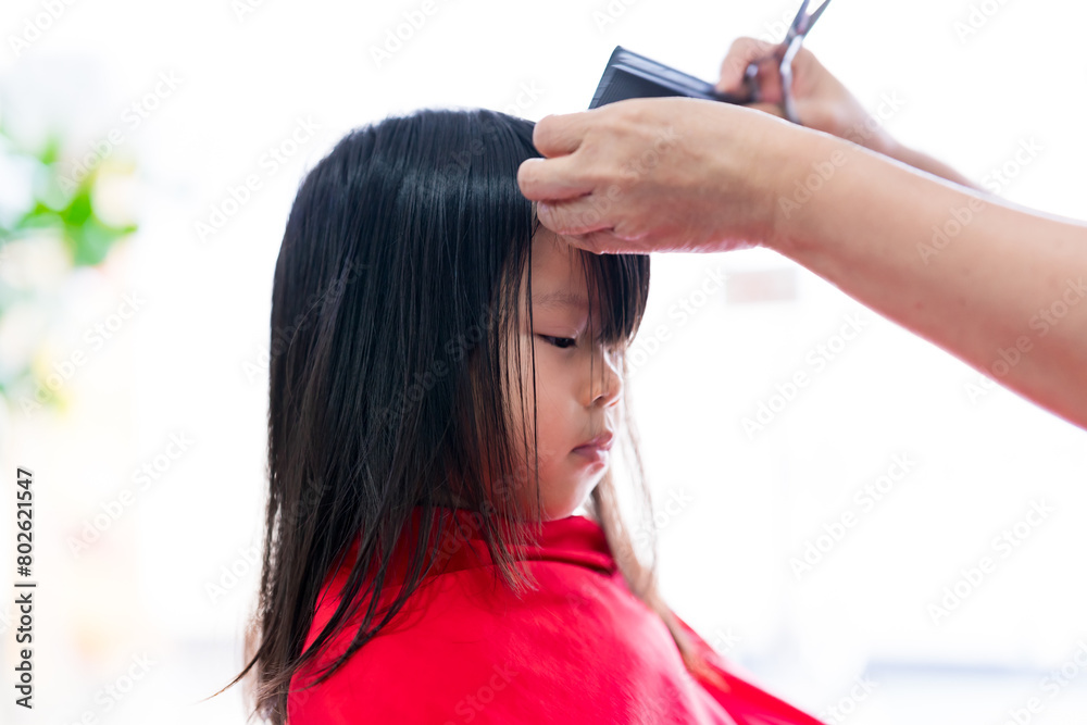 Naklejka premium Beauty Salon Styling: Woman with scissors and comb creating a fashionable hairstyle. Closeup a little child girl cutting her hairs by professional barbers salon at Barber shop.