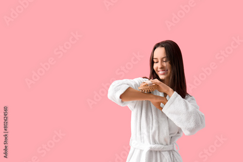 Young woman in bathrobe massaging arm on pink background