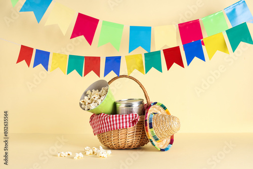 Wicker basket with popcorn, canned corn, mini sombrero and flags for Festa Junina celebration on color background photo