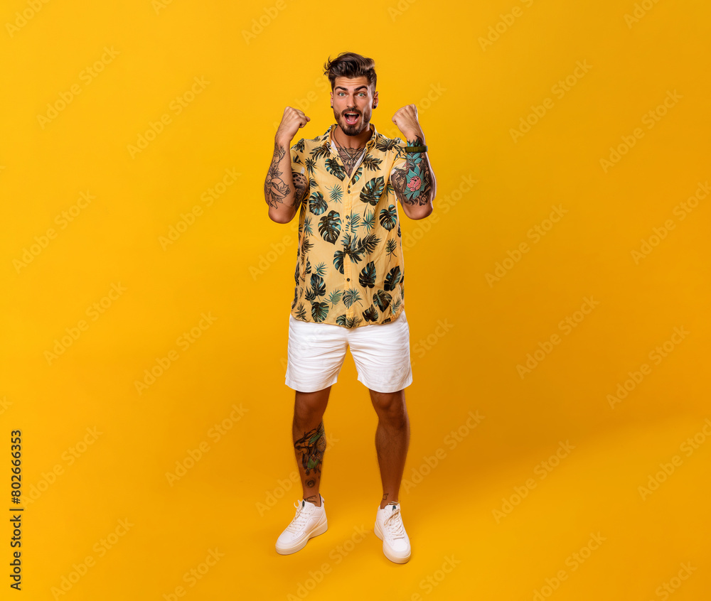 Fototapeta premium Latino man celebrating, in a yellow shirt with patterns and white shorts, tattoo in his arms, standing on a yellow background.