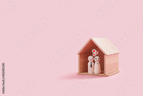 Happy wooden male and female figures inside a tiny wooden house isolated on a pink background. The couple smiling and stand close together