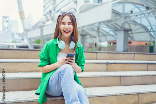 Young Asian woman using smartphone enjoying music listening through wireless headphones while sitting on steps outside in the city.