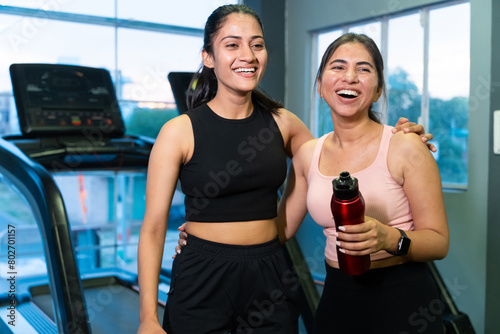 Two happy young fit indian friends women holding water bottle relaxing after workout at fitness club or gym, break time, healthy lifestyle, laughing, gossip, fun. Mental health. Stress free. 