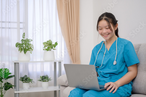 A young Asian female nurse wearing a green medical uniform works typing on a laptop sitting on a sofa in the living room at home during her break from work at a medical clinic or hospital.