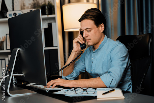 Smart businessman calling with telephone office for consulting business paragraph dynamic data marketing analysis plan surrounded paper sheet and pc monitor report at home office at night. Pecuniary.