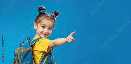 A little girl with a large school backpack on a blue background points her finger at an empty space. Copy space.