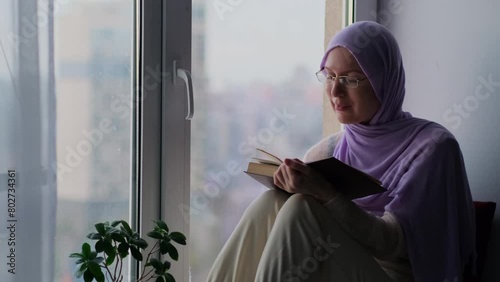 An adult Muslim woman in a hijab is reading a book while sitting at home on the windowsill with cityscape on the background. A confident woman in a headscarf is engaged in self-education. photo