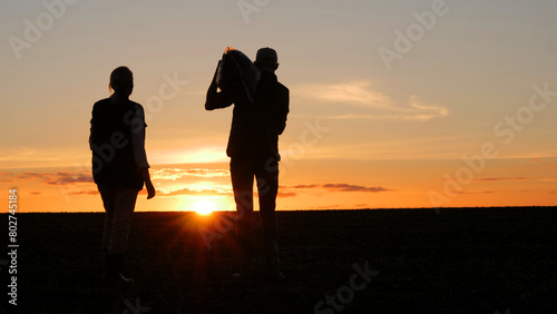 Two farmers are standing in a field at sunset. A man holds a bag on his shoulder, next to his wife