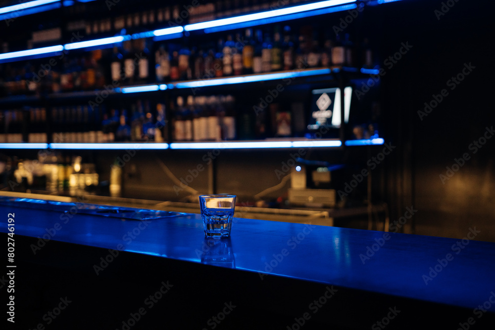 A bar with a blue counter and shelves of liquor. The bar is empty and the lights are on.