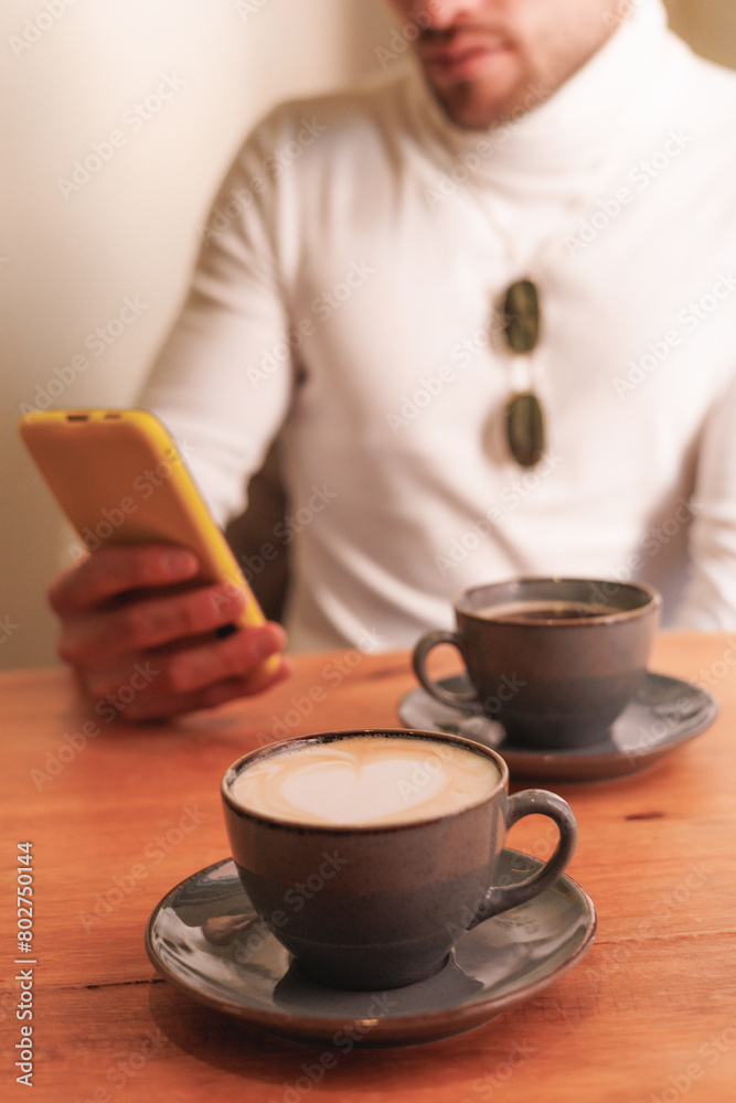 Unrecognizable latin man using cellphone in a coffee shop. Vertical view. Coffee in the foreground. Unrecognizable latin man using cellphone in a coffee shop. Vertical view. Coffee in the foreground.