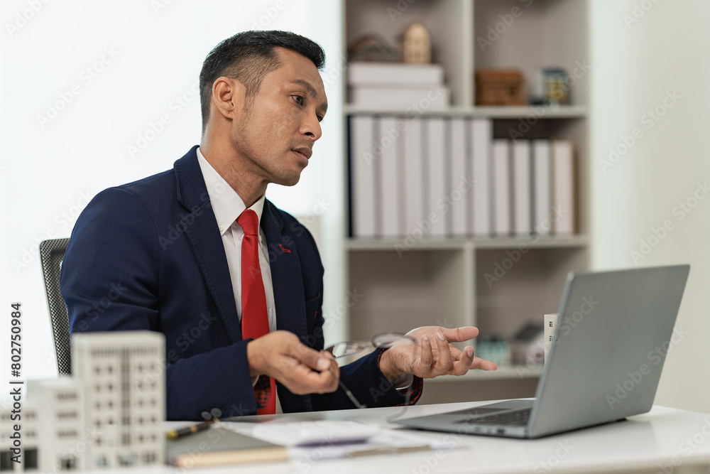 Confident professional businessman presents a client with a condominium building model offering new real estate to potential buyers via laptop online. Confident professional businessman presents a client with a condominium building model offering new real estate to potential buyers via laptop online.