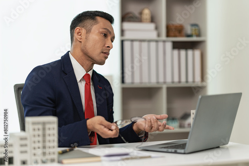 Confident professional businessman presents a client with a condominium building model offering new real estate to potential buyers via laptop online.