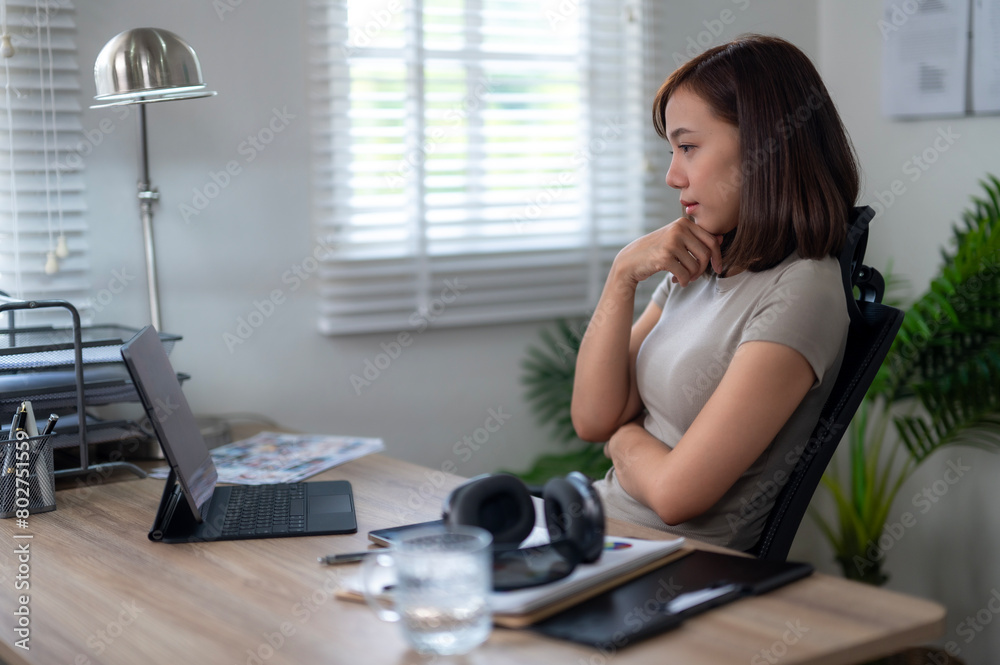 A woman sits at a desk with a laptop and a tablet. She is looking at the laptop screen and she is deep in thought. A woman sits at a desk with a laptop and a tablet. She is looking at the laptop screen and she is deep in thought.