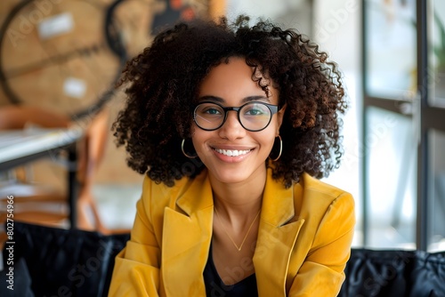 A young professional woman in a bright yellow blazer, smiling warmly while seated in a modern office environment. She wears stylish glasses and her curly hair frames her face beautifully
