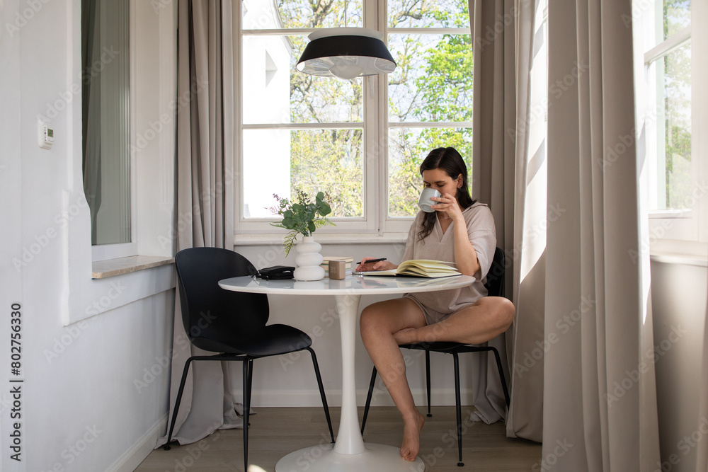 Young woman sitting at table, writing in diary and drinking coffee