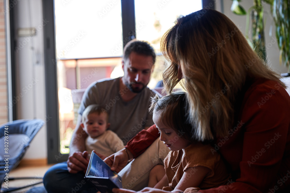 Mother and daughter reading a book while father and baby are looking them Mother and daughter reading a book while father and baby are looking them