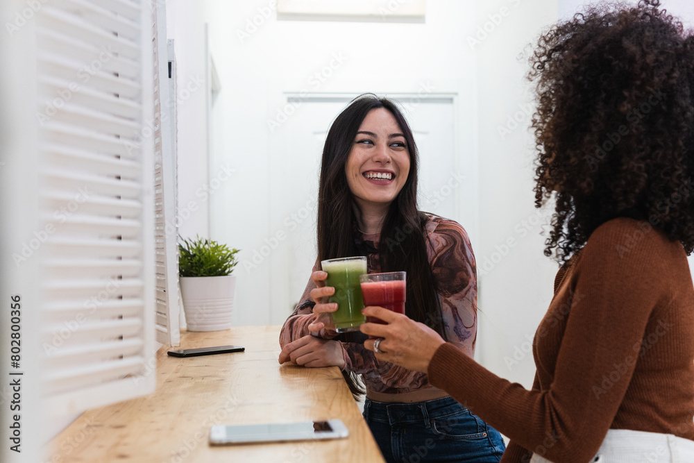 Portrait of a young women talking and laughing sitting on a coffee shop or restaurant. Two ladies smiling and looking his friend holding a smoothie. Girls having fun chatting on a bar Portrait of a young women talking and laughing sitting on a coffee shop or restaurant. Two ladies smiling and looking his friend holding a smoothie. Girls having fun chatting on a bar