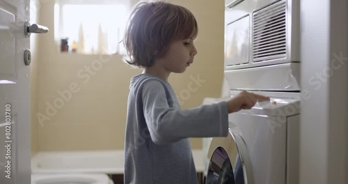 A young boy with blond hair is standing in front of a washing machine, with sunlight filtering through a window behind him. photo