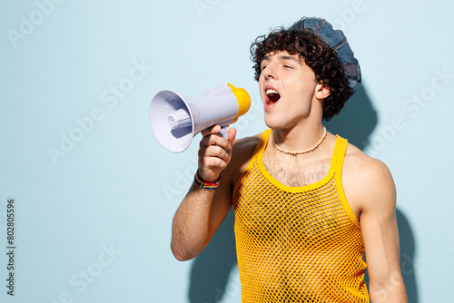 Young happy gay Latin man wear mesh tank top hat clothes scream in megaphone announces discounts sale Hurry up isolated on plain blue background studio portrait Pride day June month love LGBT concept