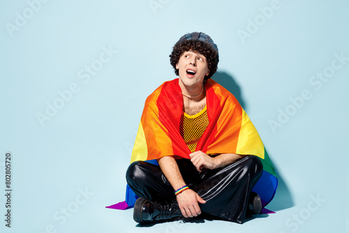 Full body young gay Latin man wear mesh tank top hat clothes wrapped in flag sits look aside on area isolated on plain pastel light blue cyan background studio. Pride day June month love LGBT concept.