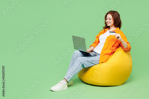 Full body young IT woman wear orange shirt t-shirt casual clothes sit in bag chair using laptop pc computer hold credit bank card shopping online isolated on plain green background. Lifestyle concept.