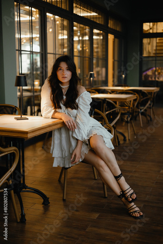 sexy young brunette wearing a light short white dress posing for a photo while sitting at a table in a street restaurant in the evening