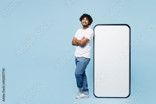 Full body side view young Indian man he wear white t-shirt casual clothes stand near big huge blank screen mobile cell phone smartphone with area isolated on plain blue background. Lifestyle concept.