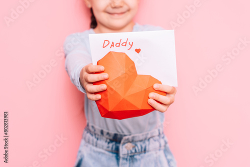 Little girl holding a drawn card and a heart for Father's Day in front of a pink background.