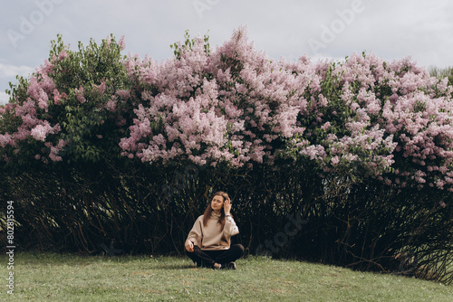 A Caucasian woman in a beige sweatshirt sits calmly near a blooming lilac, relaxing in nature, zen