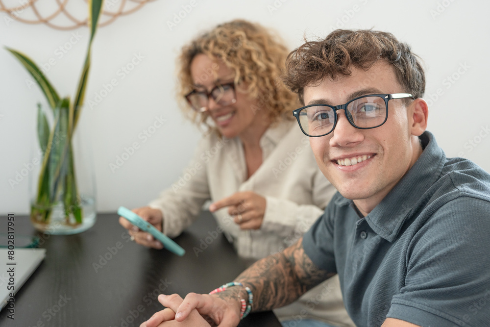 Portrait of handsome young boy looking at camera smiling sitting at table at home while blonde mom near him uses mobile phone Portrait of handsome young boy looking at camera smiling sitting at table at home while blonde mom near him uses mobile phone