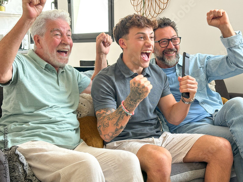 Males from multigenerational family sitting together at home looking at tv enthusiastically cheer athletes of Olympic games. Sports and participation of three attractive men of different ages